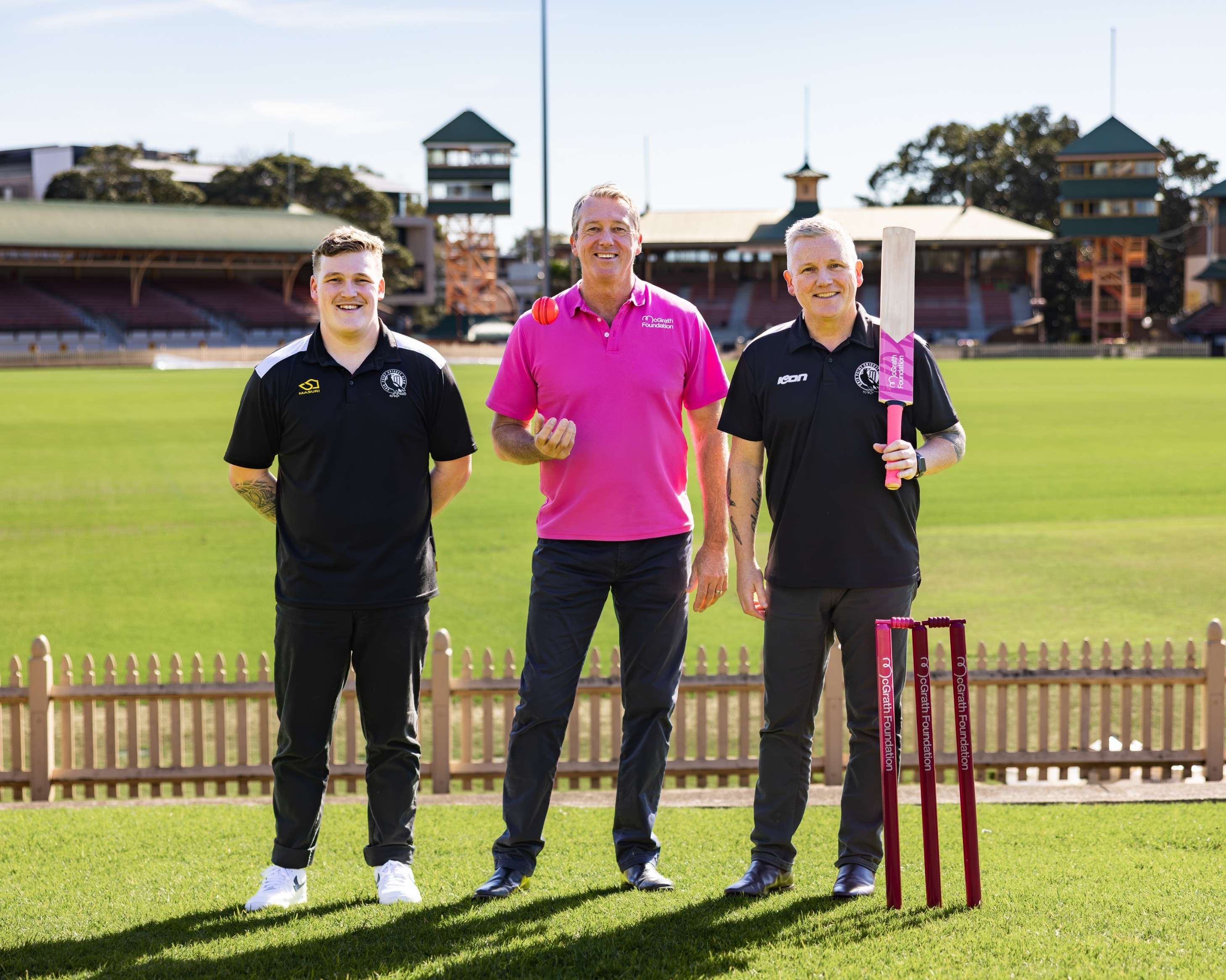 Three people stand on a cricket field at a stadium during a Pink Stumps Day promotion. One person in a pink McGrath Foundation polo shirt tosses a pink cricket ball, while another holds a cricket bat beside pink stumps branded with the McGrath Foundation logo.