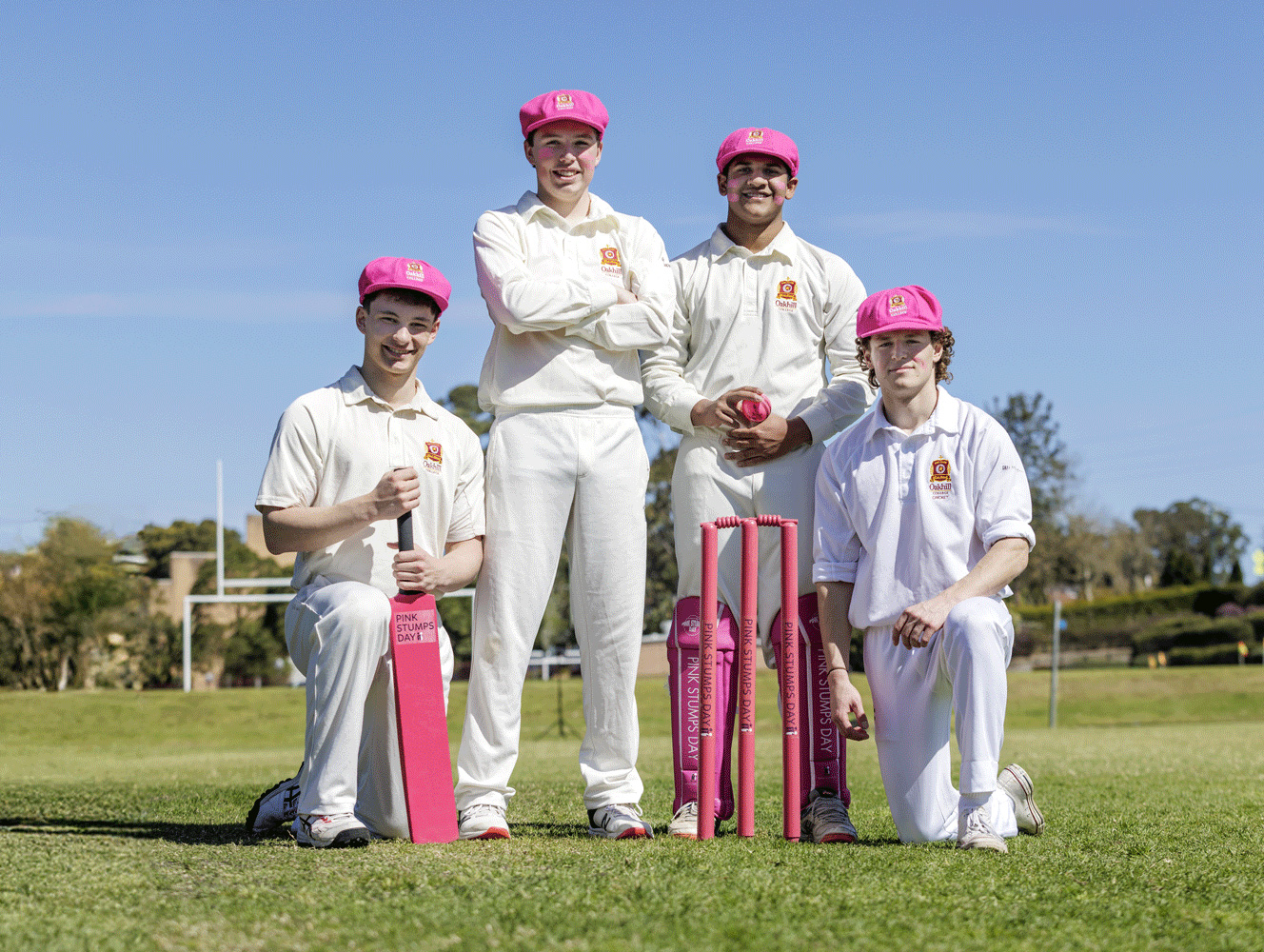 Four young cricketers in white uniforms wearing pink caps pose on a cricket field beside pink stumps and a pink cricket bat during a Pink Stumps Day event.