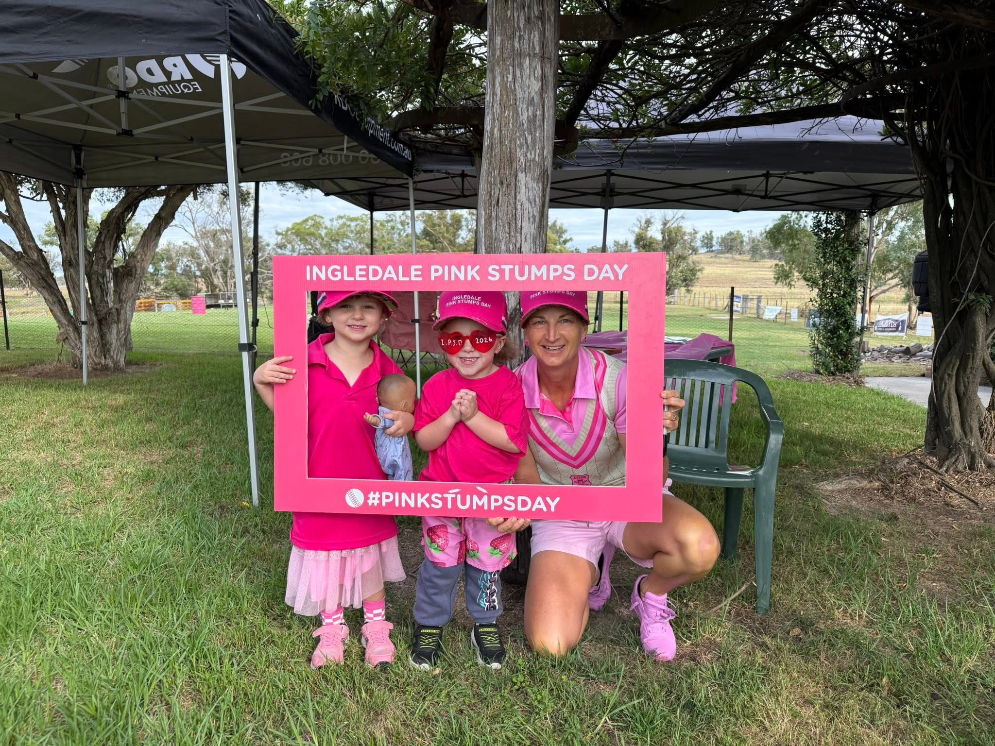 Two children and an adult dressed in pink pose behind a Pink Stumps Day photo frame at Pink Stumps Day event.