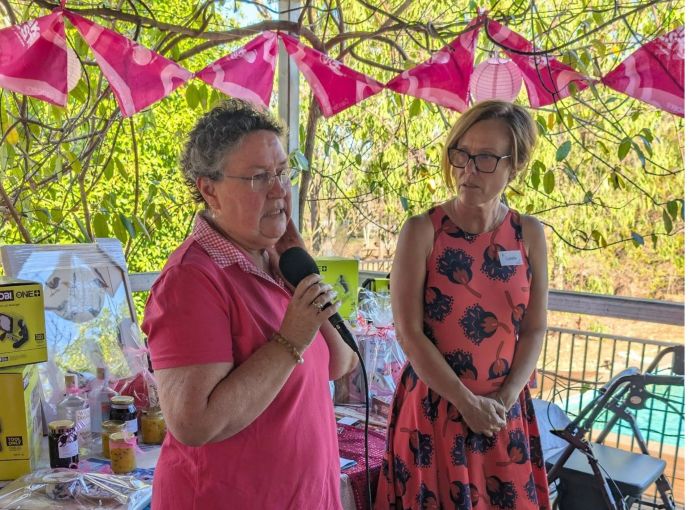 A person speaks into a handheld microphone beside another attendee on a decorated outdoor veranda. Prize hampers and donated items are displayed on a table behind them, with pink bunting hanging overhead.