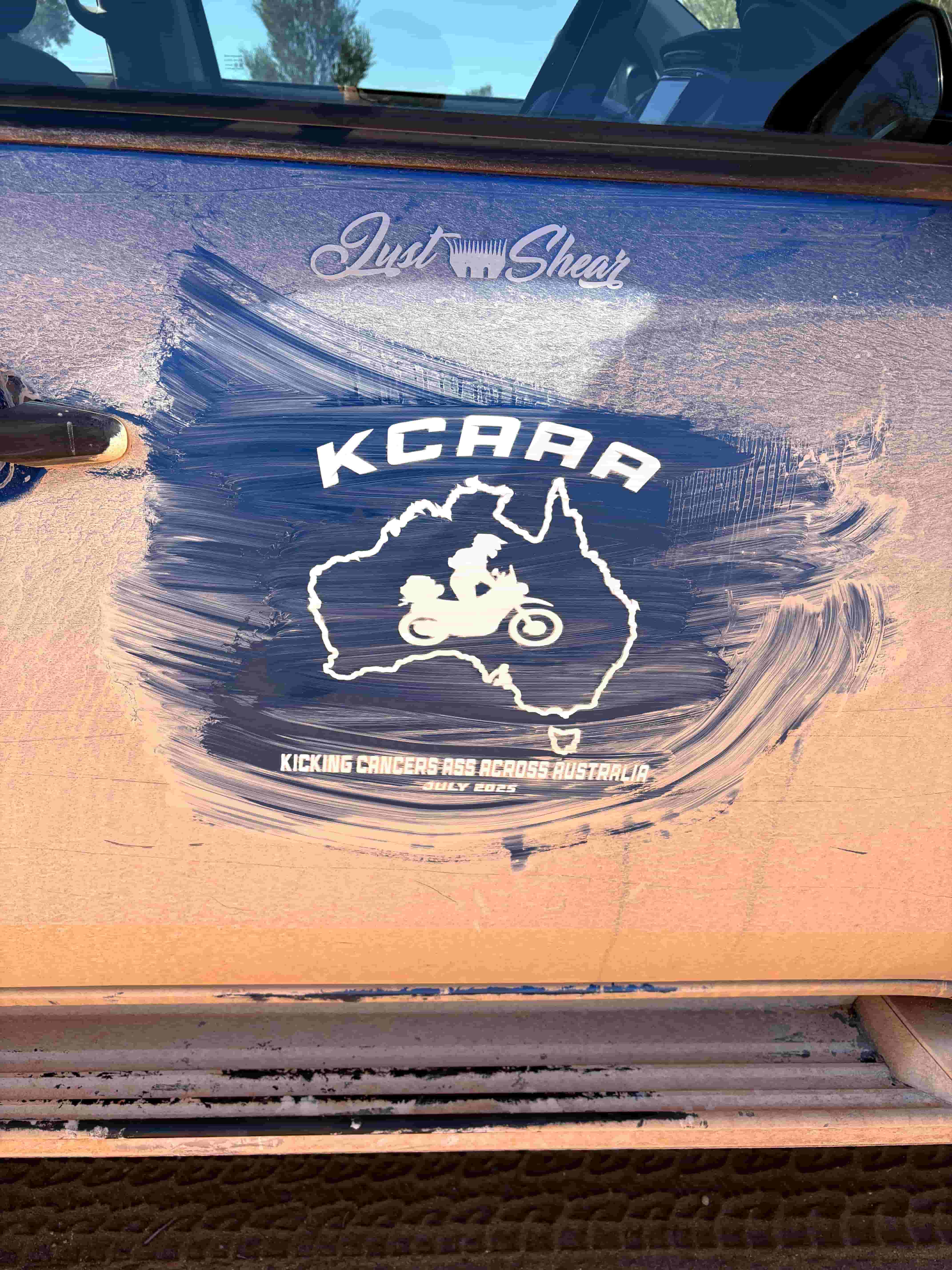 Dust-covered vehicle door displaying the KCAAA logo and the text “Kicking Cancer's Ass Across Australia – July 2025