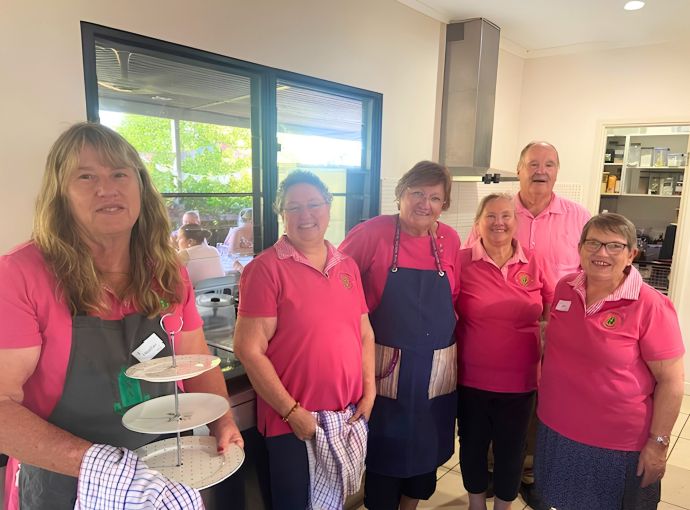 Volunteers in a kitchen at a community high tea venue, gathered for a group photo while preparing and serving food and refreshments.