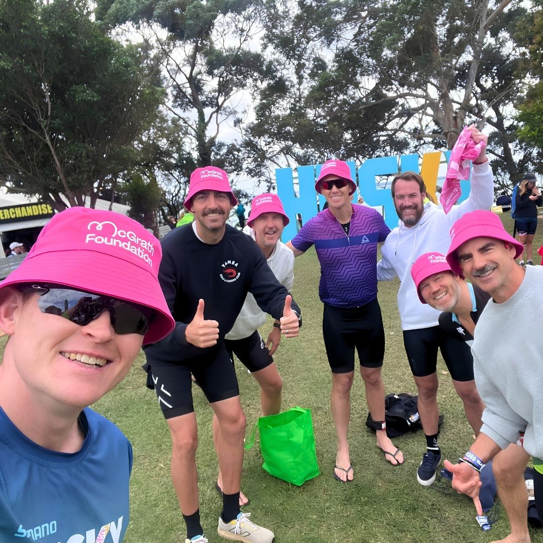 Fun run participants wearing pink McGrath Foundation hats pose together outdoors at a fundraising event, with event signage visible in the background.