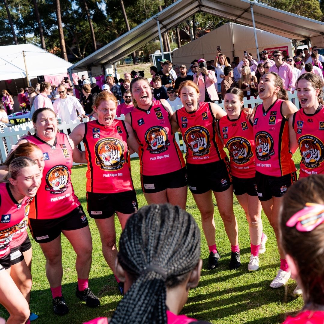Participants wearing pink football jerseys stand in a circle on a sports field during a McGrath Foundation fundraising event, with spectators gathered behind them.