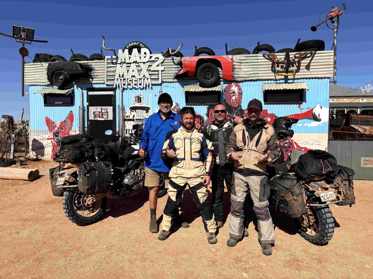 Four riders posing with adventure motorcycles outside the Mad Max 2 Museum, a corrugated metal building decorated with vehicle parts and Mad Max-themed artwork.