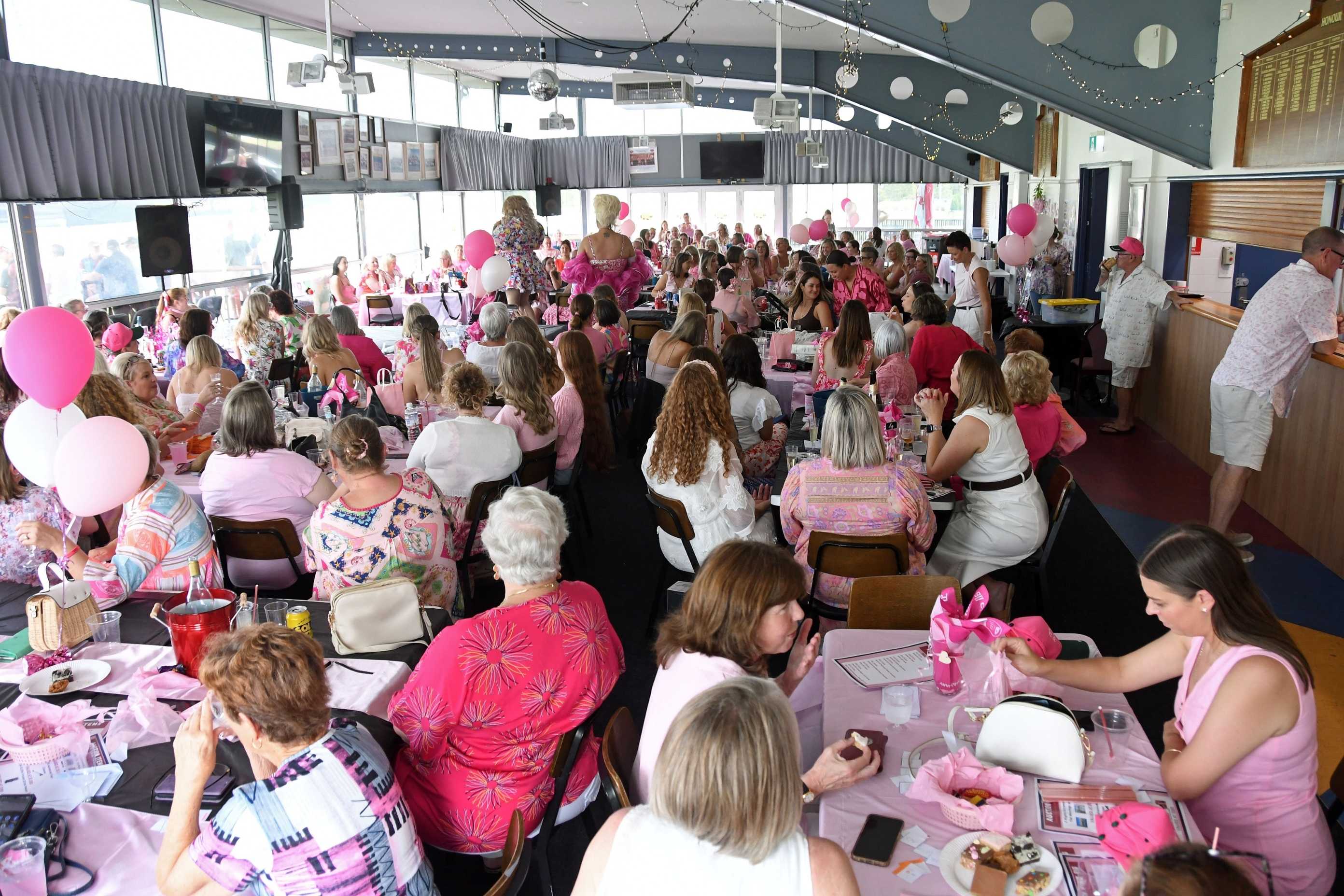 Large crowd seated at tables decorated in pink inside a function venue, attending a community fundraising event with pink balloons and themed decorations.