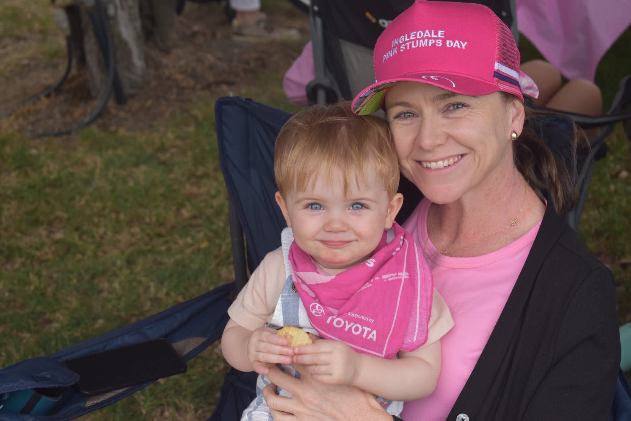 Adult wearing a pink Pink Stumps Day cap sits outdoors holding a young child wearing a pink event bandana during a community fundraising event.