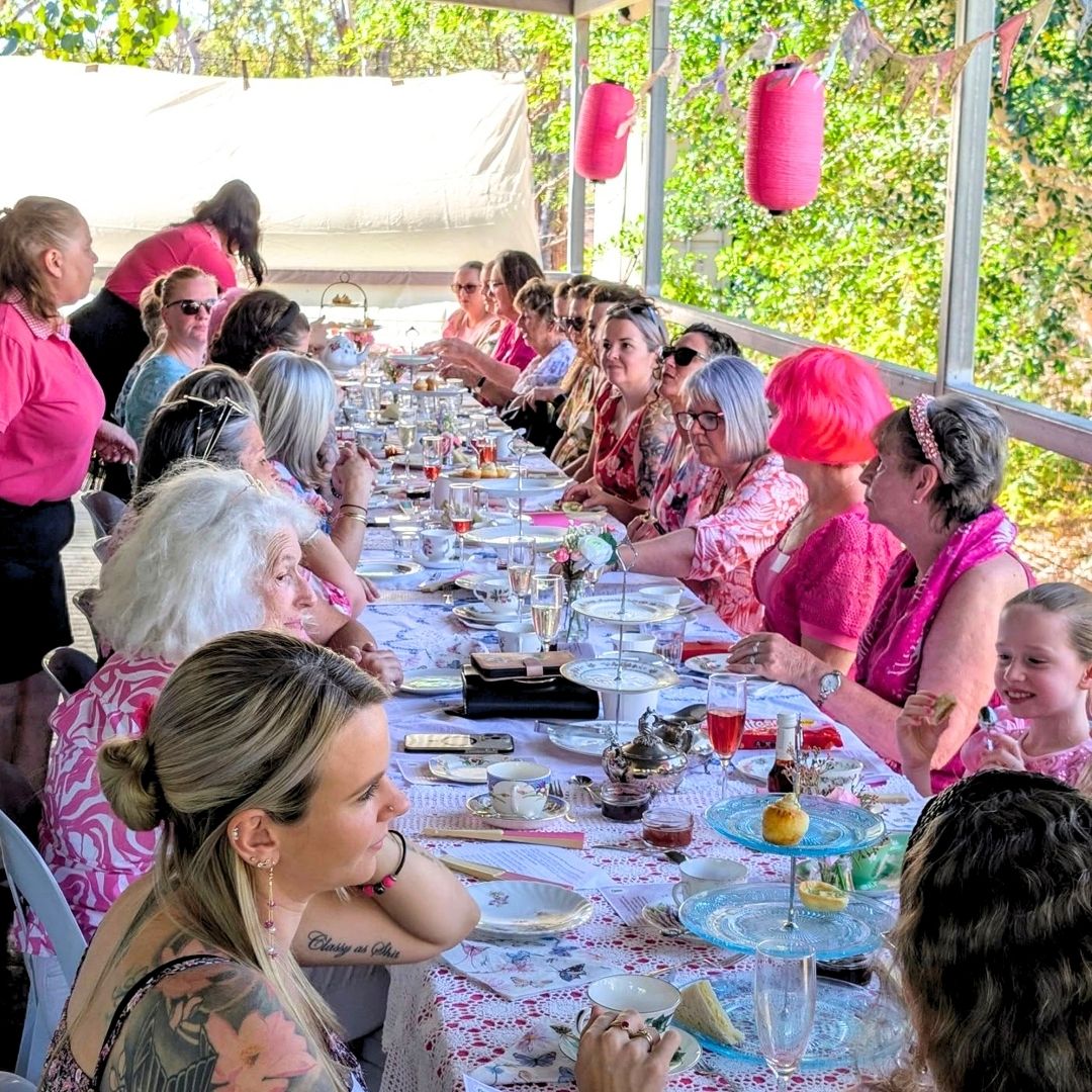 Guests dressed in pink gather around a long table for a McGrath Foundation fundraising breakfast, sharing food and conversation in an outdoor venue decorated with pink lanterns.