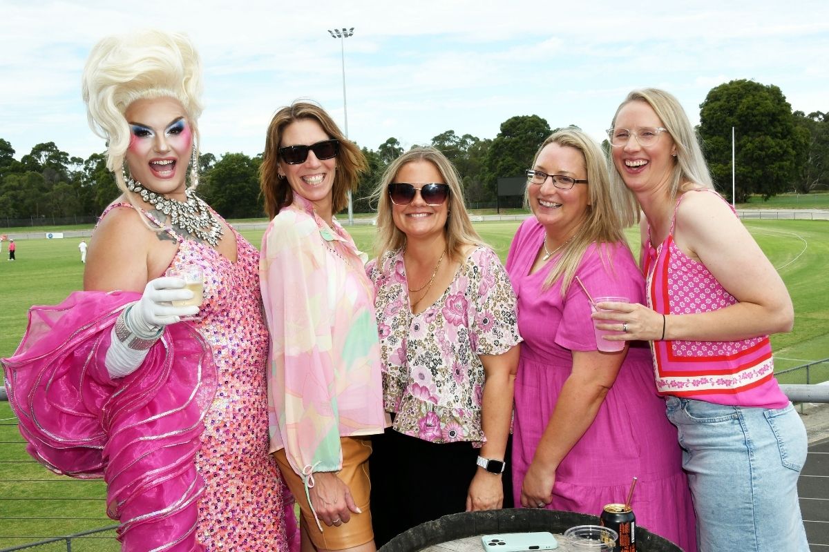 Group of people wearing pink clothing and accessories, posing together with drinks at a community sporting ground during a Pink Stumps Day event.