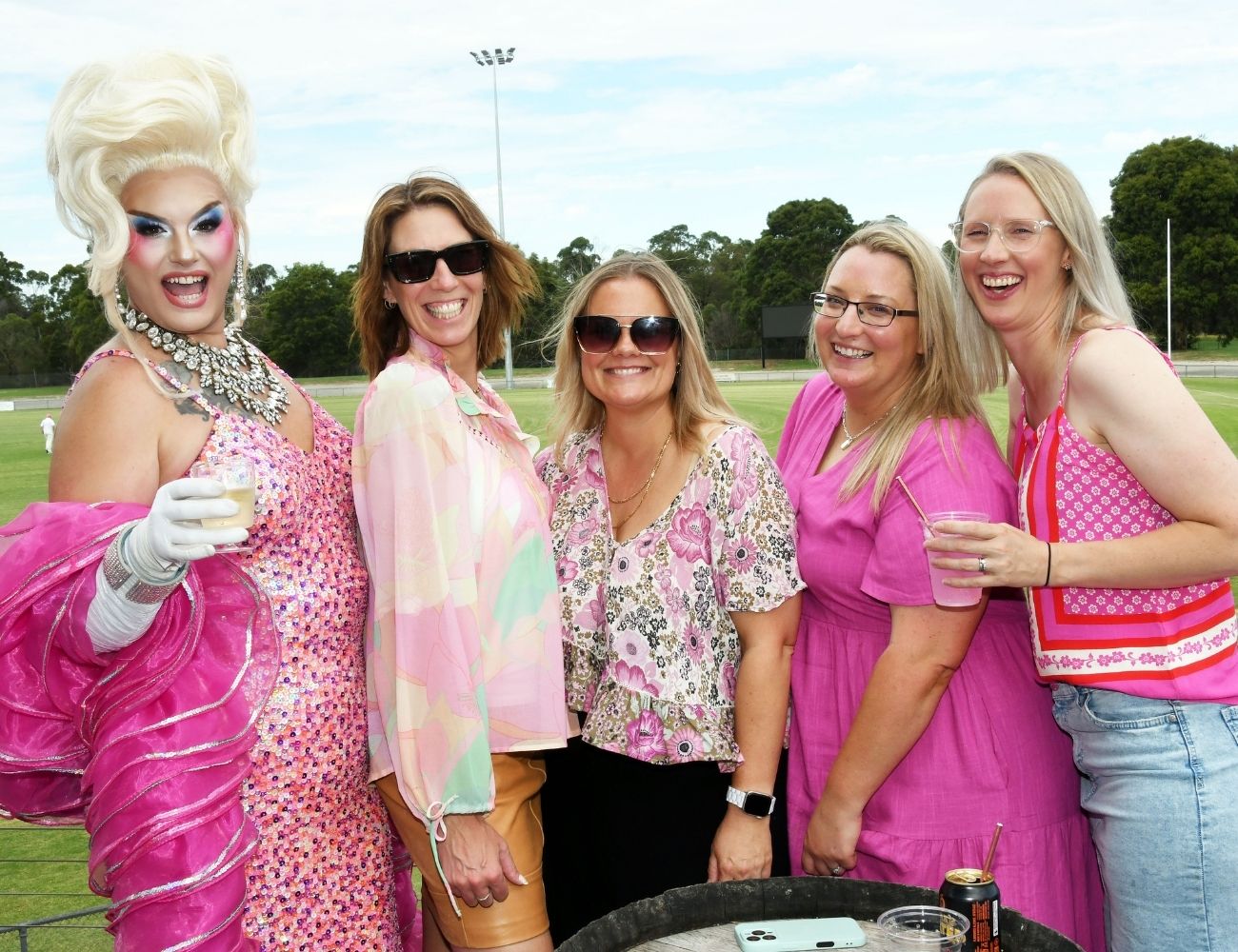 Group of people wearing pink clothing and accessories, posing together with drinks at a community sporting ground during a Pink Stumps Day event.