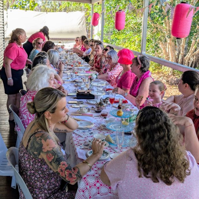Participants enjoying a community high tea event on a covered veranda, seated at a long table set with tea service, refreshments, and pink-themed decorations.
