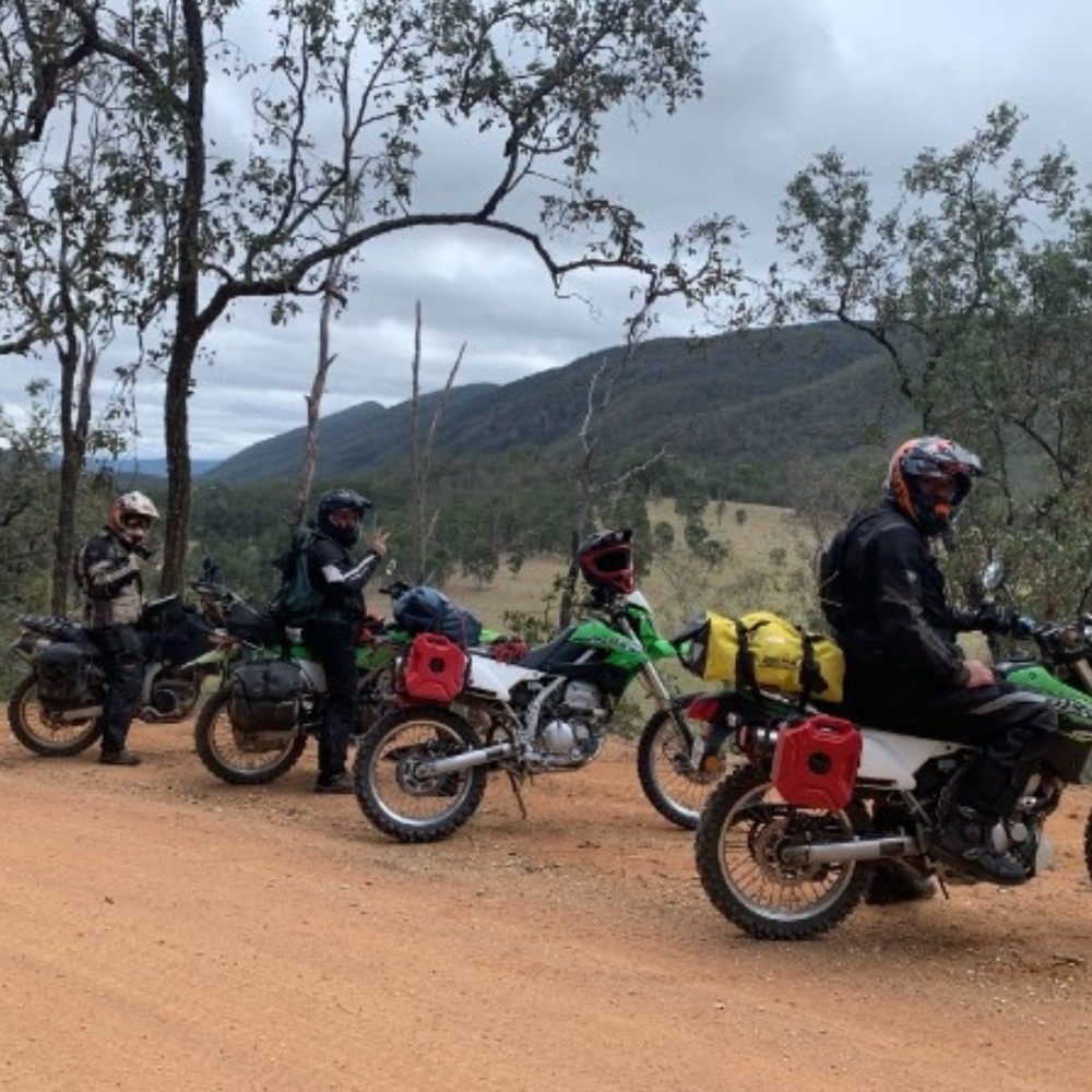 Three adventure motorcycles loaded with camping gear are parked on a dirt road overlooking a bushland valley and mountain range under a cloudy sky.